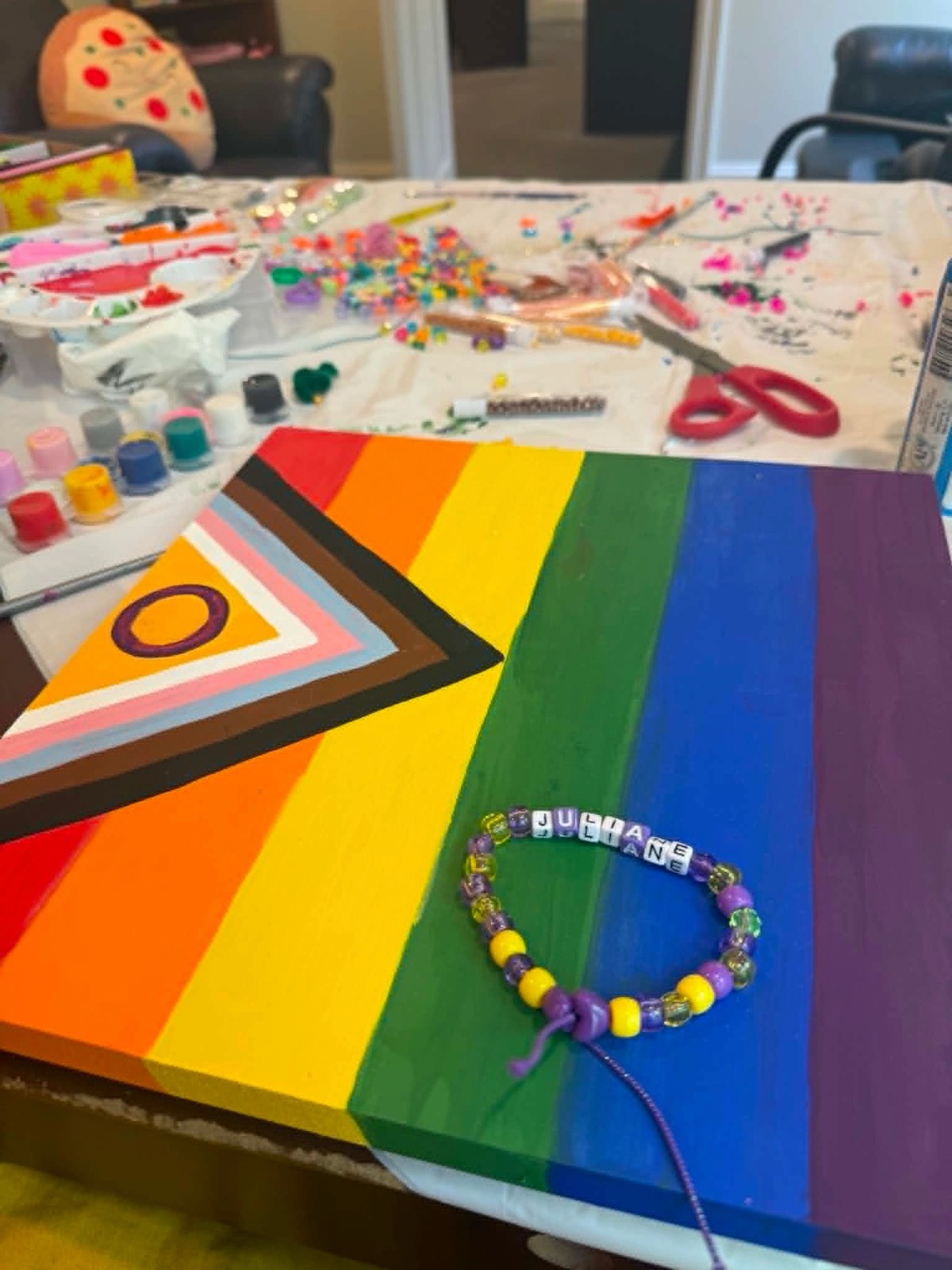 A craft table with a hand-painted Progress Pride flag and a beaded bracelet spelling Juliane